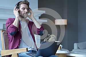 Young german guy sitting on wicker chair with laptop on his lap listening music in headphones