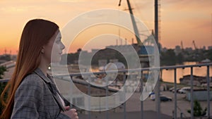 Young ginger woman in jacket walking across bridge, industrial factory background, sunset