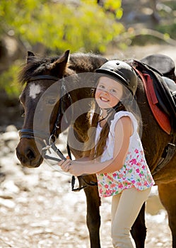 Young girl 7 or 8 years old holding bridle of little pony horse smiling happy wearing safety jockey helmet in summer holiday