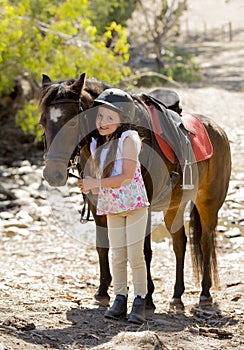 Young girl 7 or 8 years old holding bridle of little pony horse smiling happy wearing safety jockey helmet in summer holiday