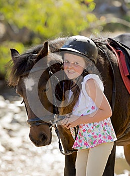 Young girl 7 or 8 years old holding bridle of little pony horse smiling happy wearing safety jockey helmet in summer holiday