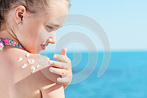 Young girl applyng sun protector cream at her shoulder on the beach close to tropical turquoise sea under blue sky