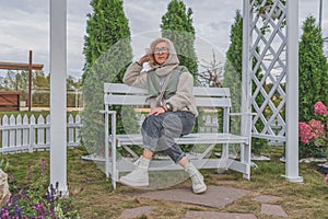 Young girl in autumn clothes is sitting in gazebo on bench in public recreation park. Looking away.