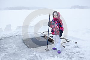 Young girl with axe in hand for ice cutting is standing near ice-hole at lake