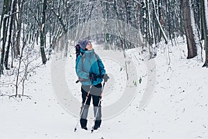 Young girl with backpack and trekking poles looks aside hiking through the winter forest