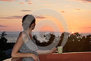 Young girl on balcony with cup of coffee looking at the sea and the sunset