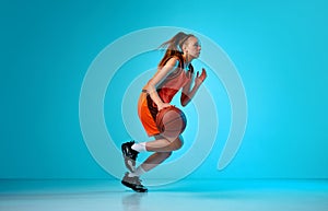 Young girl, basketball player in motion, running with ball against blue studio background in neon light