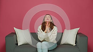 Young girl begging, pleading folded hands asking help or forgiveness, sitting on sofa on pink studio background