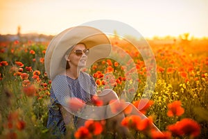 Young girl in a big hat sitting on a poppy field in sunset
