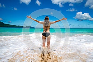 Young girl in bikini with raised arms greeting tropical sea and sun, on beach, freedom, vacation