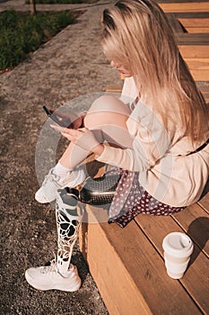 A young girl with a bionic prosthesis sits and looks at a smartphone in the park.