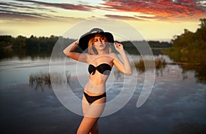 A young girl in a black bikini poses against the backdrop of a river and an epic crimson sky at sunset.