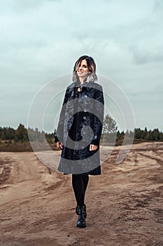 A young girl in a black coat walking on a sandy dirt road under stormy sky