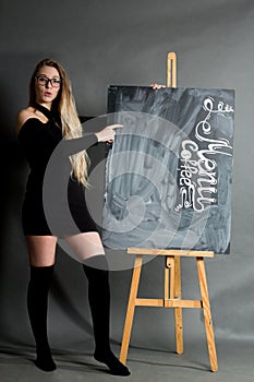 A young girl in a black dress and hetaera, with long hair and glasses, points to a chalkboard