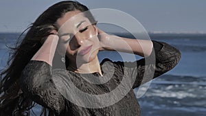 A young girl in a black dress strokes her hair, in the background ocean, sunny day