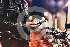 Young girl in black leather jacket using smartphone on background illumination glow bokeh light in night atmospheric city, hipster