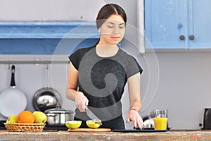 A young girl in a black t-shirt makes a fruit breakfast and cuts apple on a light wooden board in a kitchen