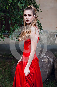 Young girl with blond flowing long hair and beautiful make-up, in a long red dress, stands on the street