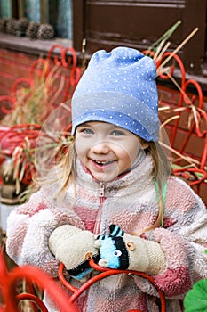 A young girl with blonde hair smiles in a blue hat with white polka dots, a colorful jacket, and white and pink gloves.