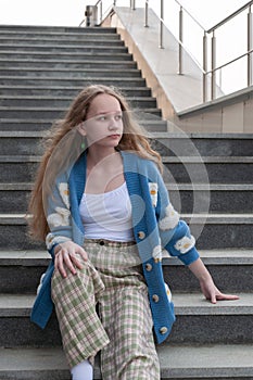 A young girl, blonde with long hair, in a blue cardigan is sitting on the steps of a gray marble staircase