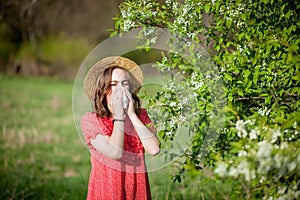 Young girl blowing nose and sneezing in tissue in front of blooming tree. Seasonal allergens affecting people. Beautiful lady has