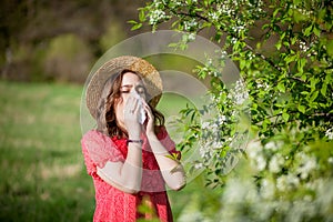 Young girl blowing nose and sneezing in tissue in front of blooming tree. Seasonal allergens affecting people. Beautiful lady has