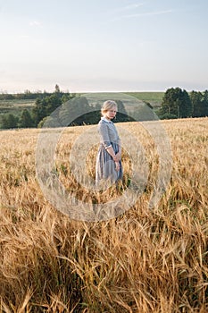 Young girl in blue dress with collected hair looks at camera in field of ears at sunrise. Green boscage in background