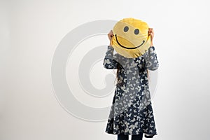 Young girl in blue dress hide her face with yellow smili pillow isolated over white background