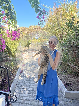 A young girl in a blue dress and sunglasses is holding a beautiful gray tabby cat in nature, Cyprus. A disgruntled cat tolerates