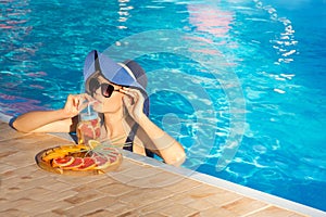 Young girl in a blue hat relaxes in the pool and drinks uit