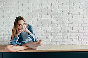 Young girl in a blue shirt sits cross-legged against a large white brick wall and looks thoughtfully off to the side with a smile