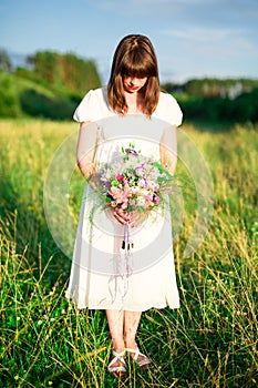 Young girl with bouquet in white dress stands in the middle of the field with his head down. Sadness, loneliness