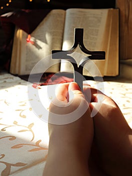 A young girl or a boy holds a cross in hands and prays. The background is an open old Bible book decorated with a red cloth with b