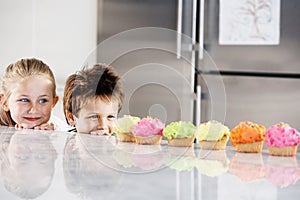 Young girl and boy peeking over counter at row of cupcakes