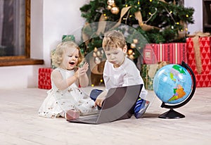 Young girl and boy watching something on laptop computer beside Christmas tree. Globe next to kids