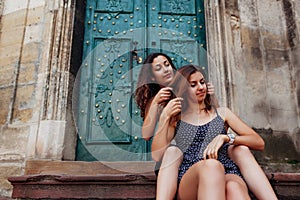 Young girl braids her best friend`s hair sitting on stairs outdoors. Happy women having fun