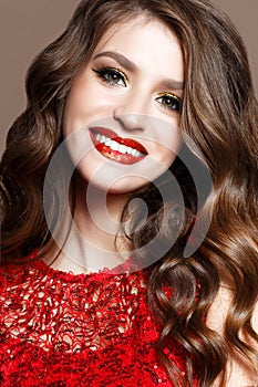 A young girl in a brilliant red dress and evening makeup. Beautiful smiling model in a New Year`s image with curls. Shining skin,