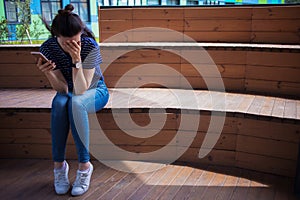 Young girl, brunette with long hair, with a phone in her hand, is crying, covering face with hands, on a wooden stairs.