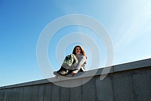 A young girl in camouflage pants and berets sits on a high granite stone fence against a blue sky.