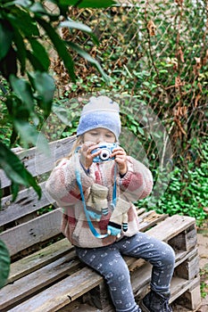 Young girl capturing moments with a blue camera on a wooden bench in a picturesque park.