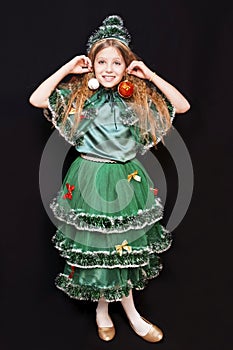 Young girl in a carnival costume of a Christmas tree on a black background holds a toy for a Christmas tree and smiles