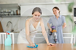 Young girl cleaning the table surface, mother vacuum-cleaning in the kitchen