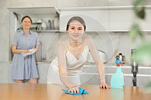 Young girl cleaning the table surface, mother wiping plates in the kitchen