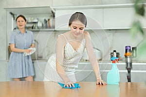 Young girl cleaning the table surface, mother wiping plates in the kitchen