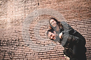 Young girl climb on the back of her boyfriend, piggyback, in front of a brick wall having fun and laughing loudly