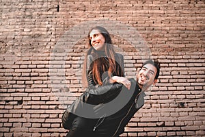 Young girl climb on the back of her boyfriend, piggyback, in front of a brick wall having fun and laughing loudly