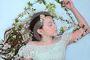 A young girl with closed eyes lies with blooming cherry branches in her long hair on a turquoise background. Portrait