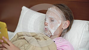 Young girl with cloth cosmetics mask on skin face lying on bad. Teenager girl using smartphone while cosmetics mask in