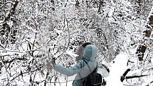 Young girl in coat going through branches in wilderness snowy forest. Female with backpack walking at snow woodland