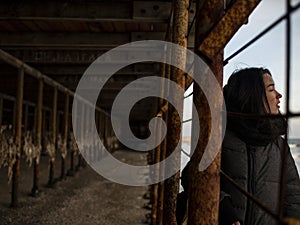 Young girl in a coat stands near metal rusty structures near the sea coast